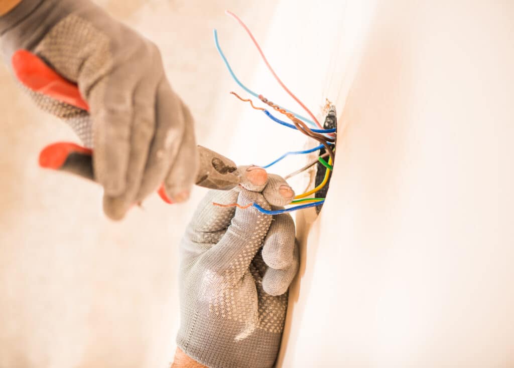 Close view of an electrician's gloved hands as they work with wires through a hole cut in a white wall.